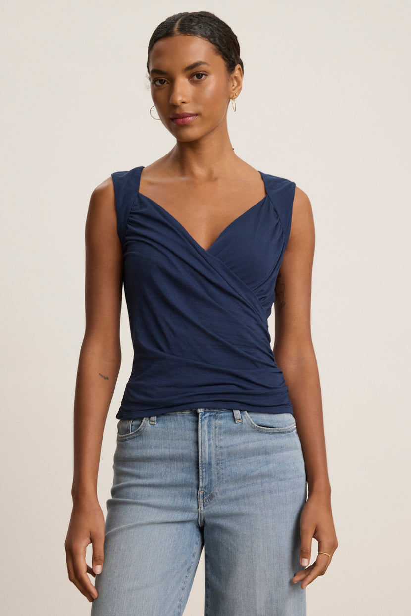 A woman stands against a plain light background wearing the Velvet by Graham & Spencer AZURA TANK TOP, a sleeveless navy blue wrap with a romantic front crossover, paired with light blue jeans.