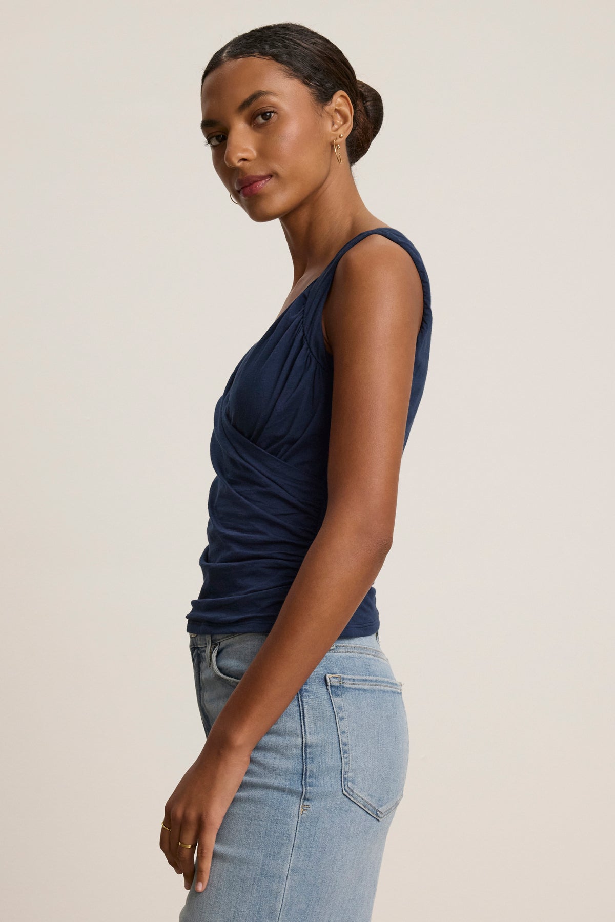 A woman stands sideways against a plain light background, wearing the AZURA TANK TOP by Velvet by Graham & Spencer—a navy sleeveless, romantic crossover wrap style—paired with light blue jeans.-39119703638209