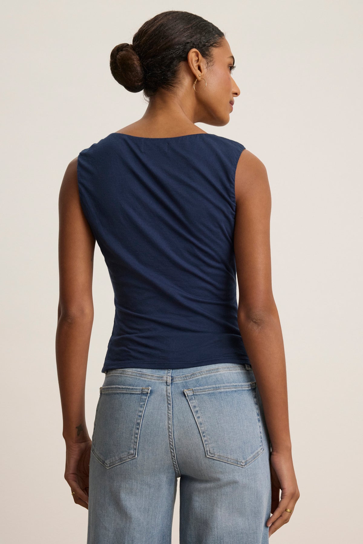   A woman stands facing away, wearing the AZURA TANK TOP by Velvet by Graham & Spencer in navy with light blue jeans, her hair pulled back in a neat bun. 
