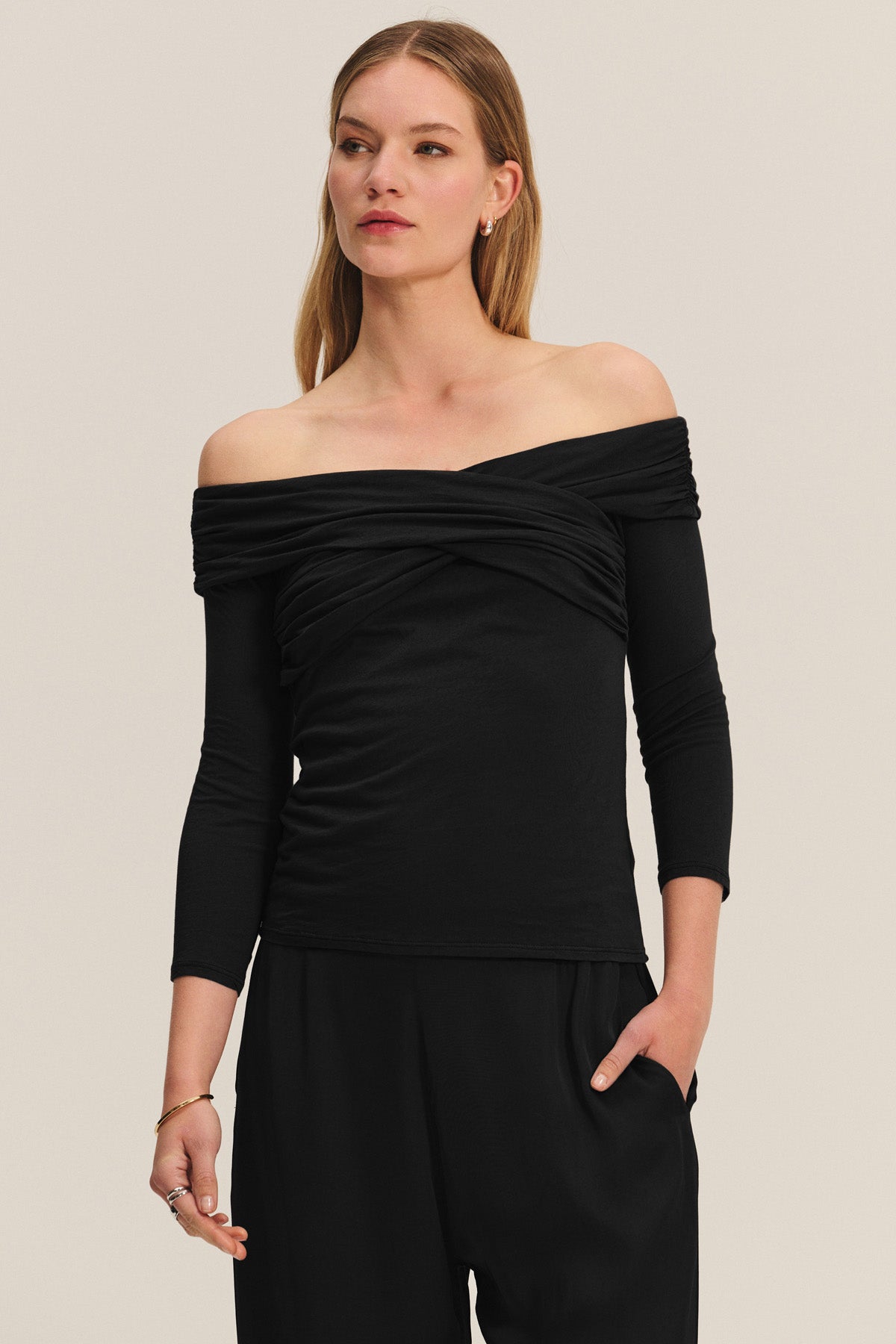 A woman wears the Velvet by Graham & Spencer CLARISSA TEE, an off-the-shoulder long-sleeve black top, paired with black pants. She stands with one hand in her pocket against a neutral background.-40688648749249