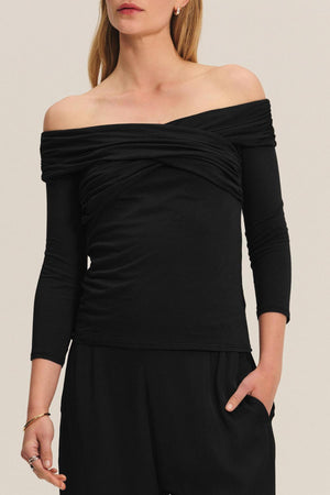A woman wearing the CLARISSA TEE by Velvet by Graham & Spencer, an off-the-shoulder black top with three-quarter sleeves, paired with black pants and standing with one hand in her pocket.