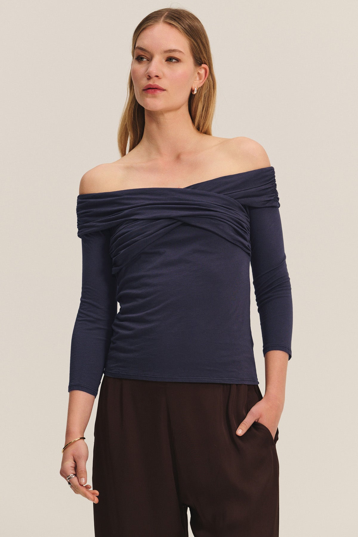 A woman wears the CLARISSA TEE by Velvet by Graham & Spencer—an off-the-shoulder, navy, three-quarter sleeve top—paired with dark brown pants, standing with one hand in her pocket.-40921836519617