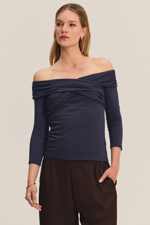 A woman wears the CLARISSA TEE by Velvet by Graham & Spencer—an off-the-shoulder, navy, three-quarter sleeve top—paired with dark brown pants, standing with one hand in her pocket.
