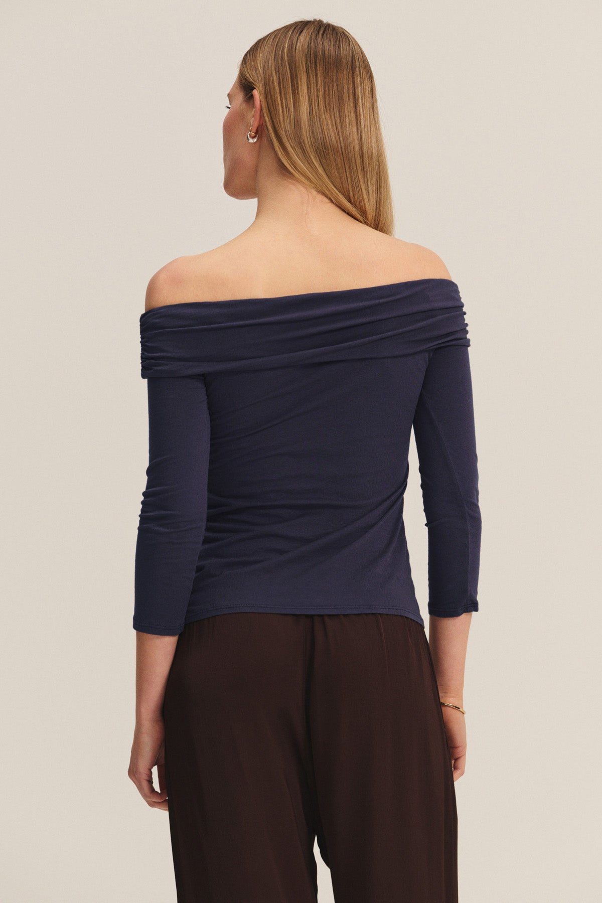 A woman faces away wearing the CLARISSA TEE by Velvet by Graham & Spencer—an off-the-shoulder navy top in gauzy whisper fabric with three-quarter sleeves—paired with loose dark pants, set against a plain light background.-40921836486849