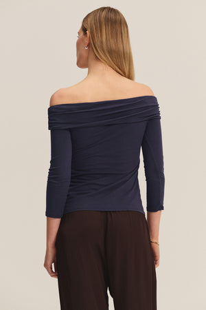 A woman faces away wearing the CLARISSA TEE by Velvet by Graham & Spencer—an off-the-shoulder navy top in gauzy whisper fabric with three-quarter sleeves—paired with loose dark pants, set against a plain light background.