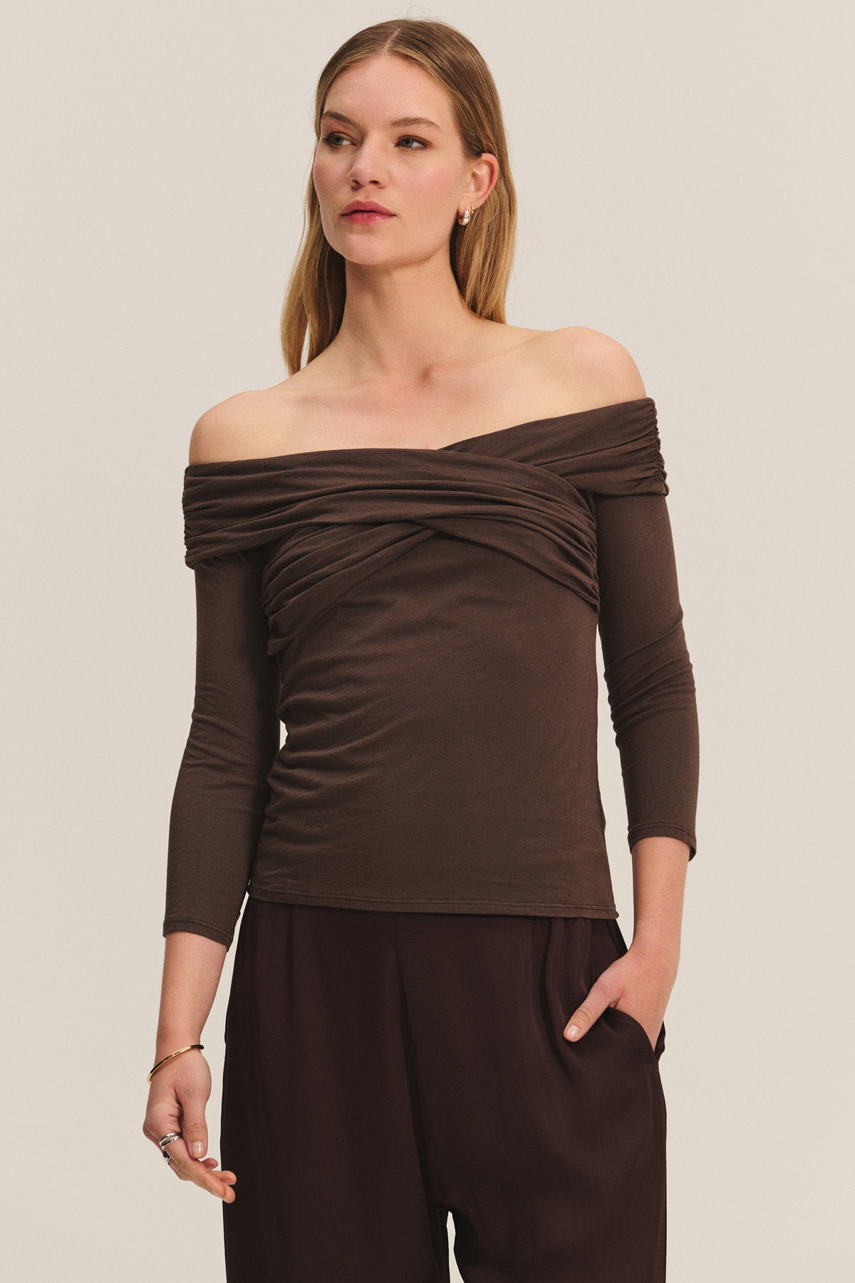 A woman wears the Velvet by Graham & Spencer CLARISSA TEE, an off-the-shoulder brown long-sleeve top, paired with brown pants, standing with one hand in her pocket against a plain background.