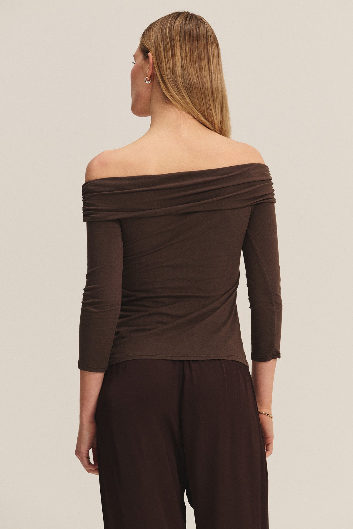 A person stands facing away, wearing the CLARISSA TEE by Velvet by Graham & Spencer—an off-the-shoulder, long-sleeve brown top—with dark brown pants against a plain background.