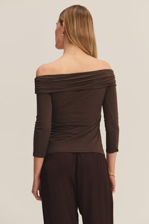 A person stands facing away, wearing the CLARISSA TEE by Velvet by Graham & Spencer—an off-the-shoulder, long-sleeve brown top—with dark brown pants against a plain background.