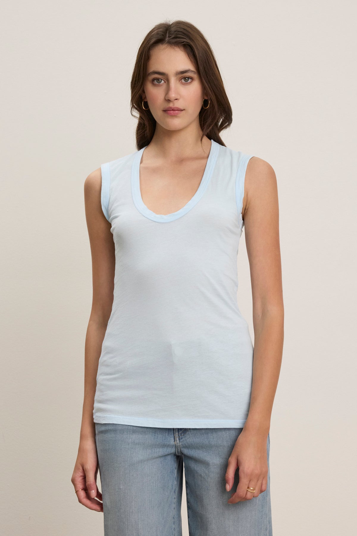 A woman faces forward in the ESTINA TANK TOP by Velvet by Graham & Spencer, a sleeveless light blue tank, paired with light blue jeans against a plain light background.-39123372572865