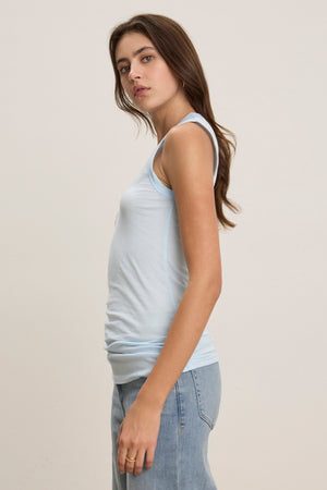 A person with long brown hair wears the Velvet by Graham & Spencer ESTINA TANK TOP in soft gauzy whisper blue and light blue jeans, standing sideways against a plain light background.