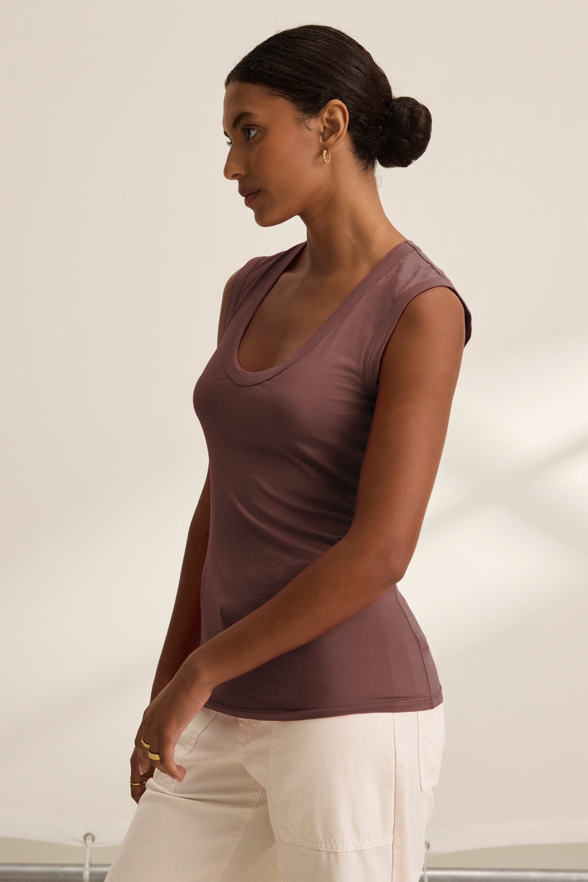 A woman with dark hair in a bun models the Velvet by Graham & Spencer ESTINA TANK TOP, featuring a scoop neck and cap sleeves in mauve, paired with light beige pants, standing in profile against a neutral background.-39520708231361