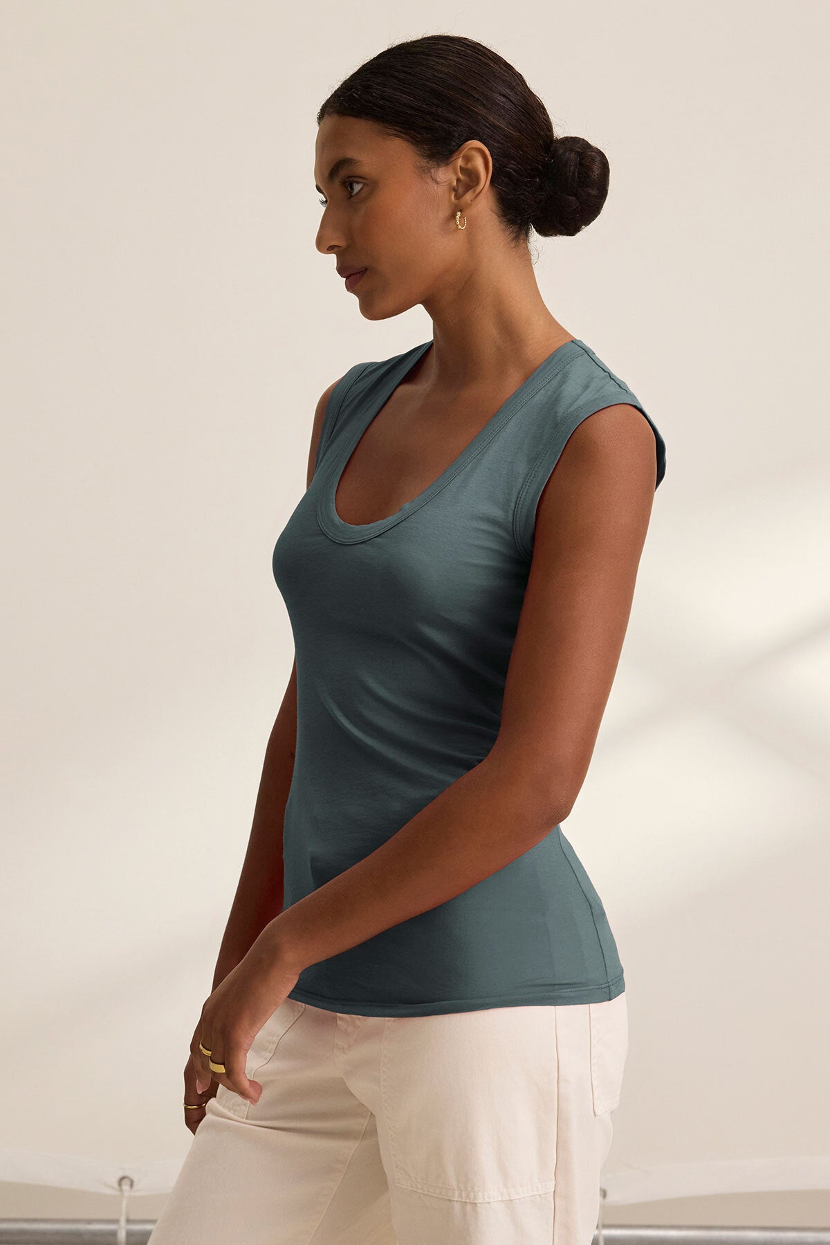   A woman stands in profile against a plain light background wearing the ESTINA TANK TOP by Velvet by Graham & Spencer and white pants. 
