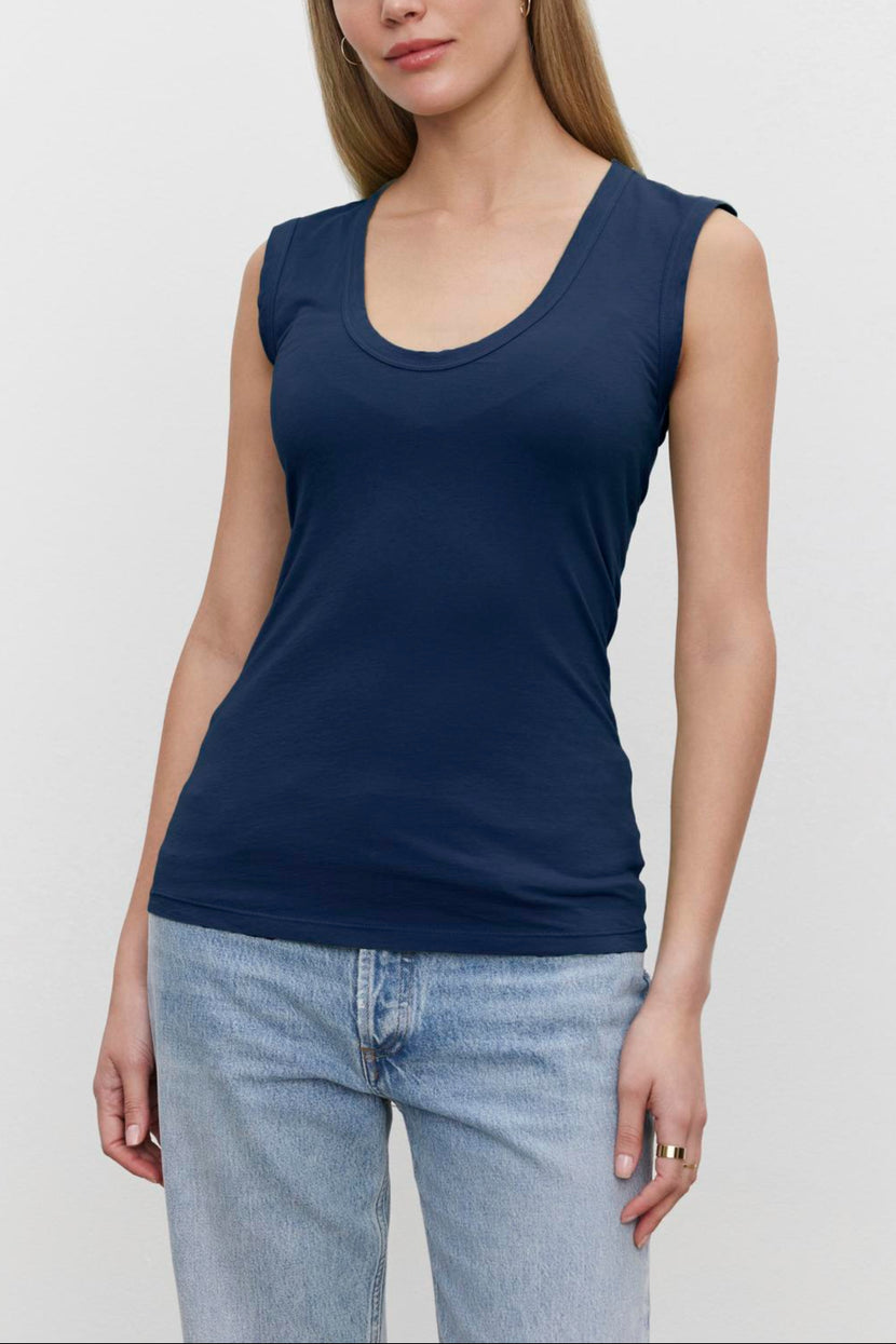 A woman with long hair is wearing a navy blue low-scoop-neck sleeveless Velvet by Graham & Spencer ESTINA TANK TOP and light blue jeans, standing against a plain white background.