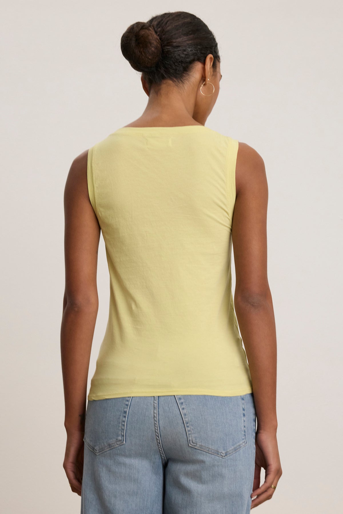 A person with their hair in a bun models the EVELYN TANK TOP by Velvet by Graham & Spencer in yellow, paired with light blue jeans, shown from the back against a plain light background.-39079878033601