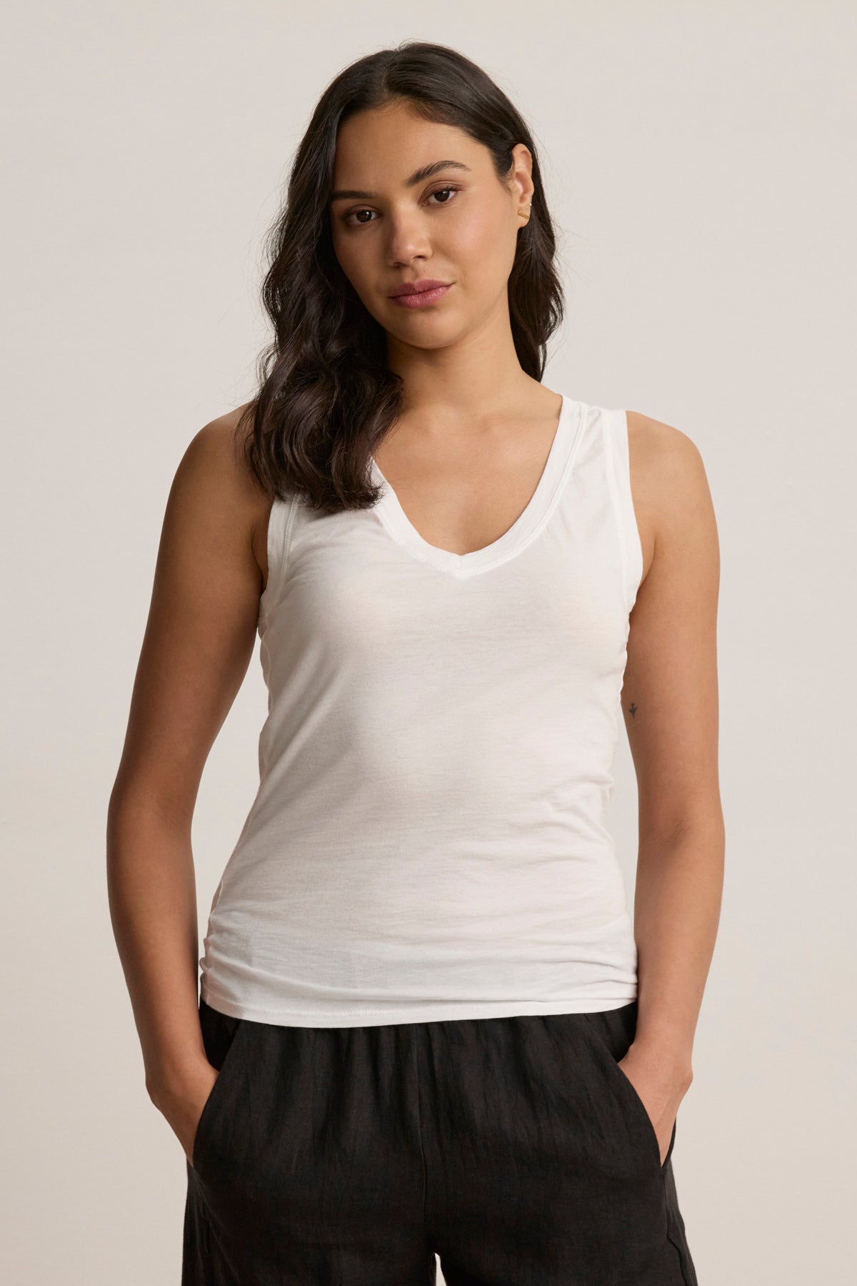 A woman with long dark hair wears the EVELYN TANK TOP by Velvet by Graham & Spencer— a versatile white sleeveless V-neck tank—paired with black pants, standing hands-in-pockets against a plain background.-39079878066369
