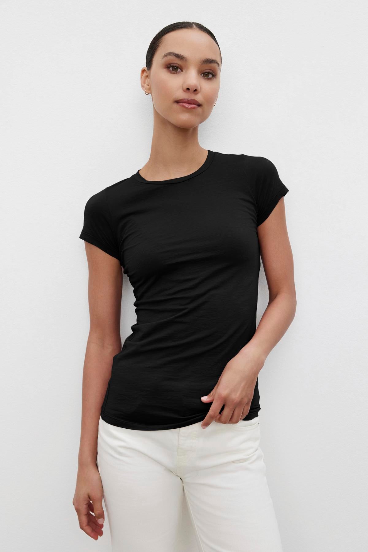 A person wearing the JEMMA TEE in a gauzy whisper black crew neck by Velvet by Graham & Spencer, paired with white pants, stands against a plain white background, facing the camera with a neutral expression.-37648662593729