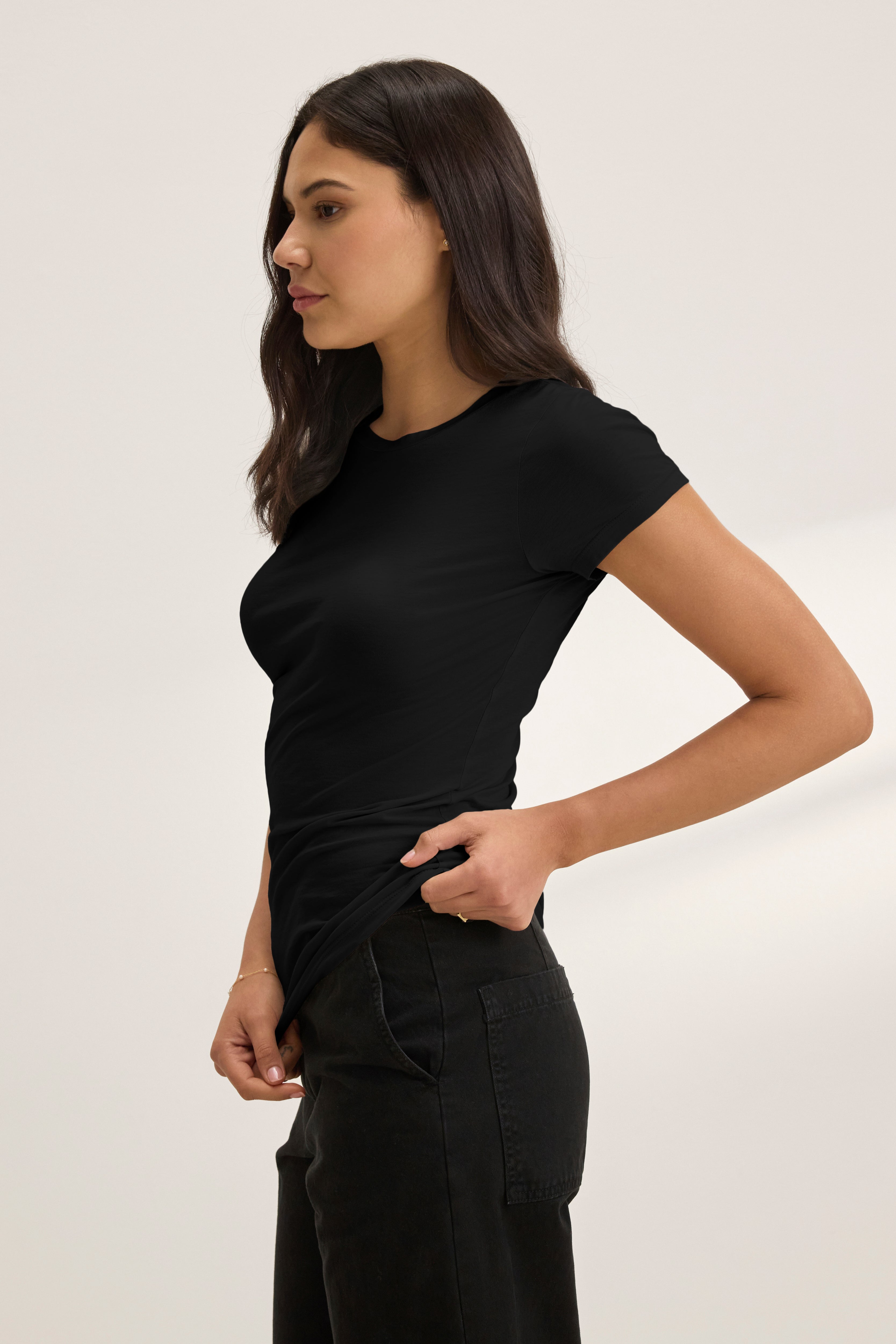   A woman stands sideways against a neutral background, wearing the Velvet by Graham & Spencer JEMMA TEE in black with dark jeans, her left hand resting on her hip. 