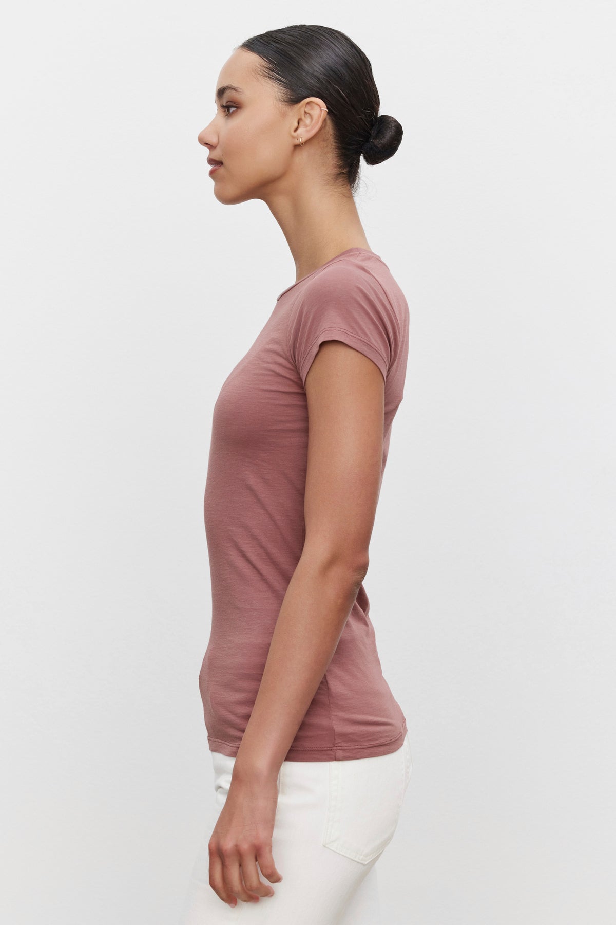   A person with tied-back hair is shown in profile wearing a crew neck Velvet by Graham & Spencer JEMMA TEE in a short-sleeve pink style and white pants, exuding the charisma of a timeless staple against a plain background. 