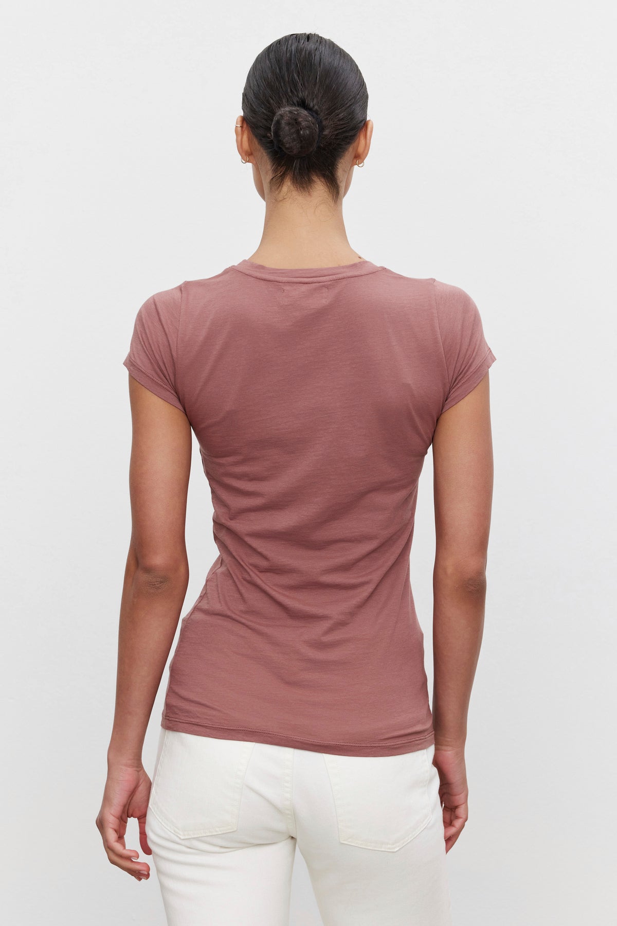   A person with dark hair in a bun is standing with their back to the camera. They are wearing an ultra-soft gauzy whisper short-sleeve, mauve-colored JEMMA TEE by Velvet by Graham & Spencer and white pants. 