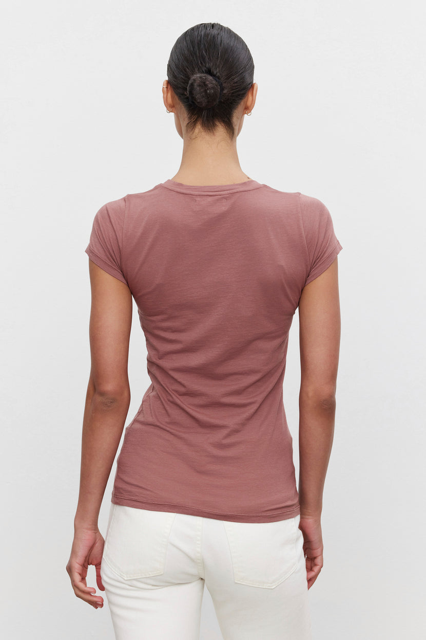 A person with dark hair in a bun is standing with their back to the camera. They are wearing an ultra-soft gauzy whisper short-sleeve, mauve-colored JEMMA TEE by Velvet by Graham & Spencer and white pants.