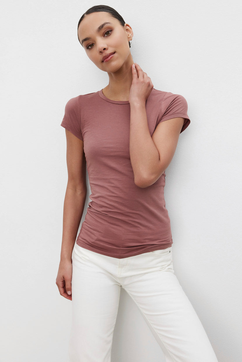 A person stands against a plain white background, wearing a JEMMA TEE by Velvet by Graham & Spencer and white pants, with one hand touching their neck.