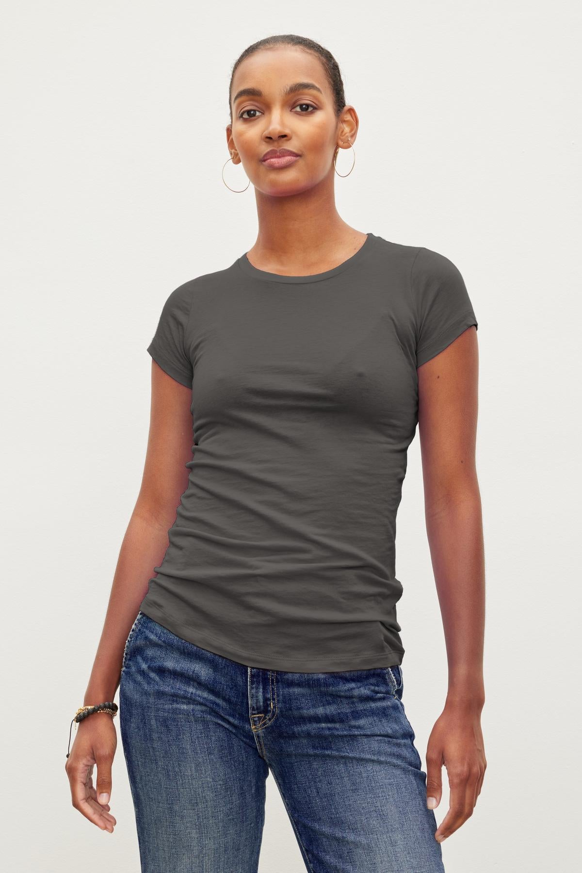   A person stands against a plain background wearing a fitted, short-sleeved gray crew neck tee and blue jeans. They have their hair pulled back and are wearing hoop earrings. The JEMMA TEE by Velvet by Graham & Spencer elevates their effortlessly casual look with its ultra-soft gauzy whisper fabric. 