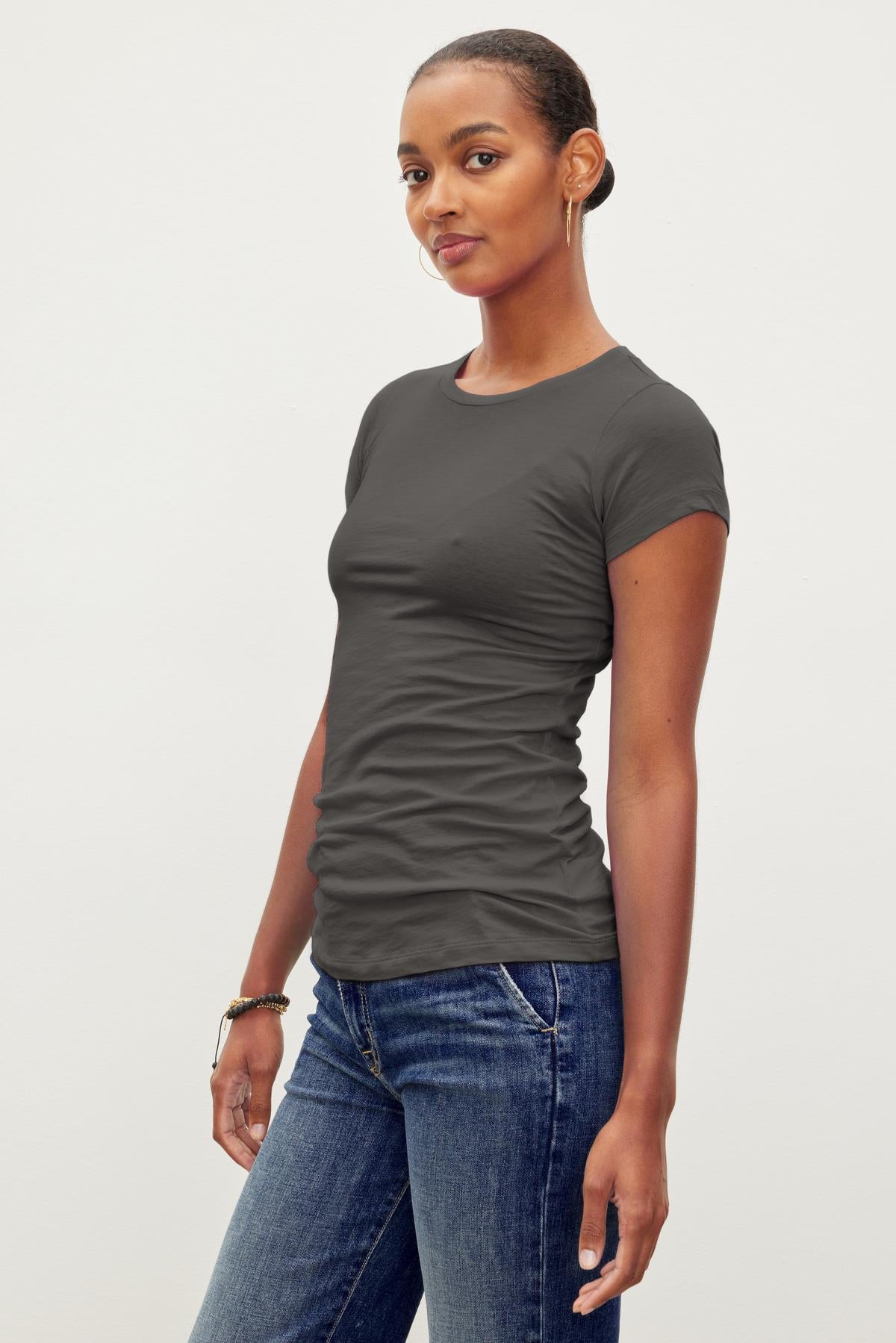 A person with short hair stands against a plain background, wearing a Velvet by Graham & Spencer JEMMA TEE in dark gray and blue jeans—a timeless staple.-37241159516353