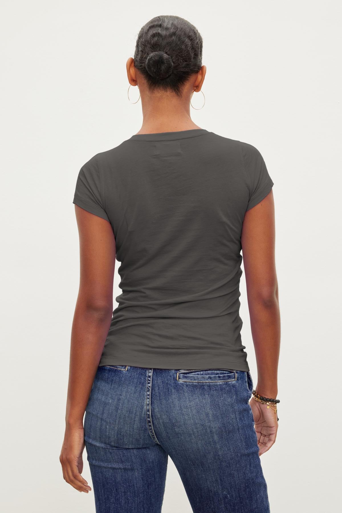   A person with a bun hairstyle is wearing a Velvet by Graham & Spencer JEMMA TEE in dark grey and blue jeans, standing with their back to the camera. 