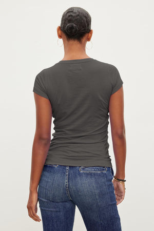 A person with a bun hairstyle is wearing a Velvet by Graham & Spencer JEMMA TEE in dark grey and blue jeans, standing with their back to the camera.