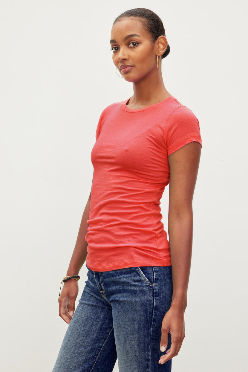 A person wearing a fitted crew neck red short-sleeve JEMMA TEE by Velvet by Graham & Spencer and blue jeans stands against a plain white background.