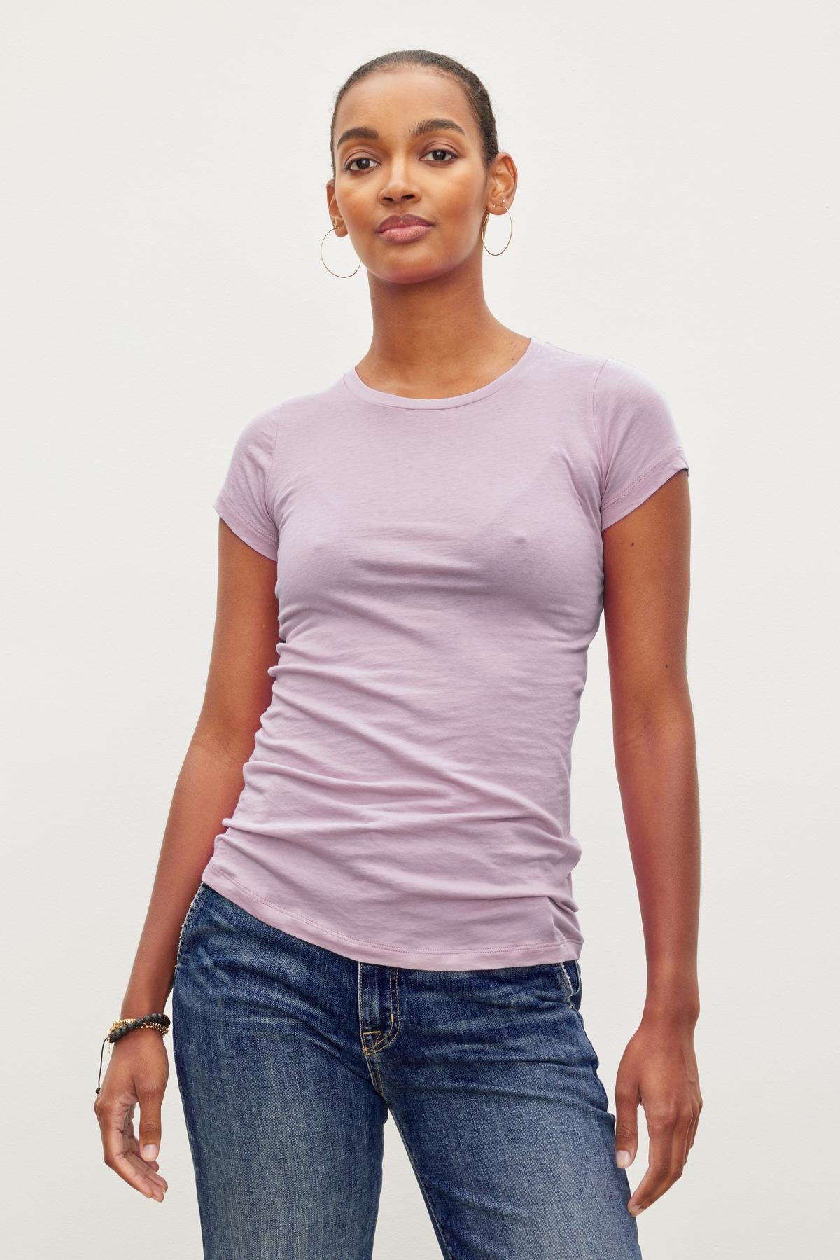   A woman wearing an ultra-soft Velvet by Graham & Spencer JEMMA TEE and blue jeans stands against a plain white background, looking straight ahead. She has hoop earrings and a bracelet on her left wrist. 