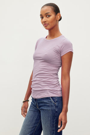 A woman with short hair wearing a light purple, ultra-soft gauzy JEMMA TEE by Velvet by Graham & Spencer and blue jeans, standing against a plain white background.