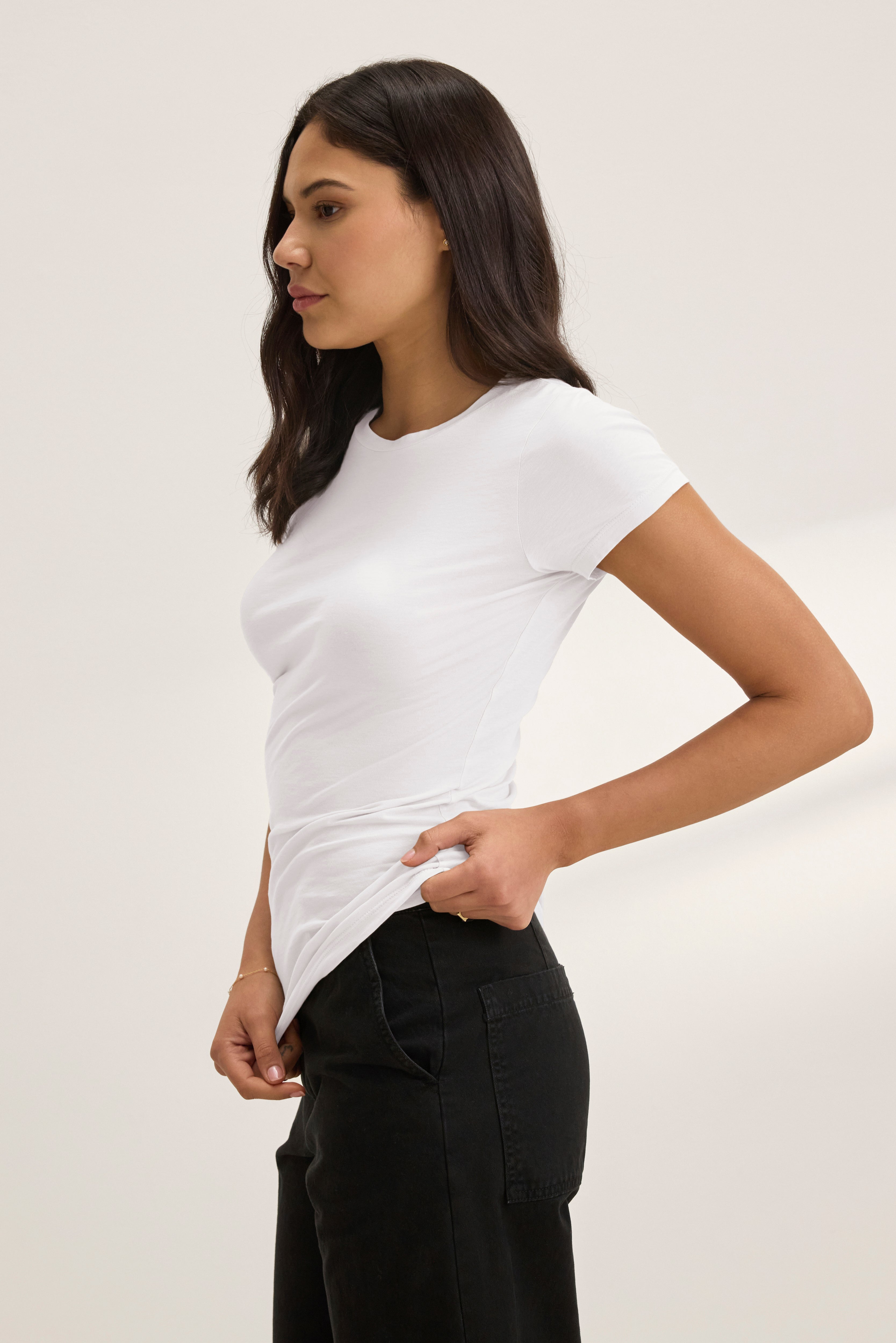   A person stands sideways against a plain light background, wearing the ultra-soft, fitted white JEMMA TEE by Velvet by Graham & Spencer and black pants. 