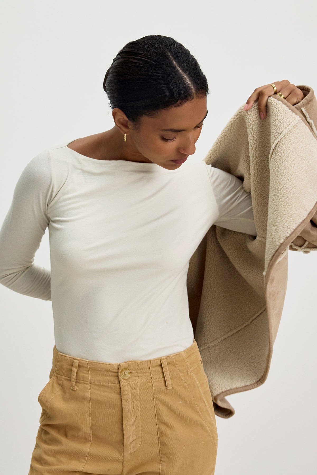   A person in a white long-sleeve shirt holds the KELLY LUXE SHERPA REVERSIBLE JACKET by Velvet by Graham & Spencer, featuring a beige faux suede finish. 