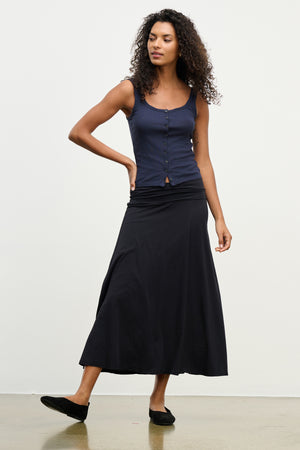 A woman poses against a plain background wearing the LUELLA SKIRT by Velvet by Graham & Spencer, paired with a sleeveless navy button-up top and black flats.