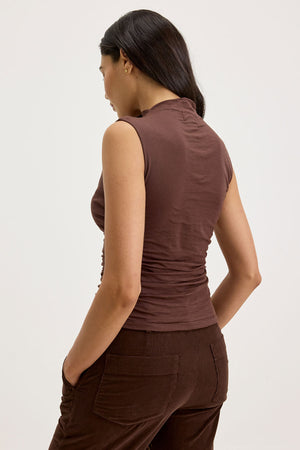 A woman with long dark hair is standing with her back slightly turned, wearing the MADELINE TANK TOP from Velvet by Graham & Spencer paired with matching brown pants. Her left hand is in her pocket, highlighting the fitted look of her outfit, while the gauzy whisper fabric adds an airiness to her ensemble.