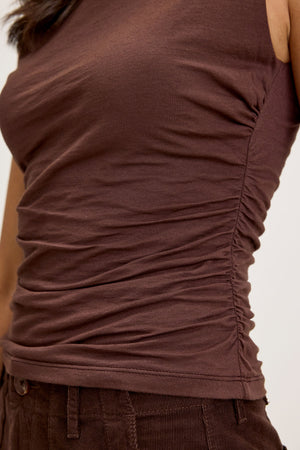 A person wearing the MADELINE TANK TOP from Velvet by Graham & Spencer, which is a brown sleeveless top with ruching on the side and a mock neck for a fitted look, paired with brown corduroy pants.