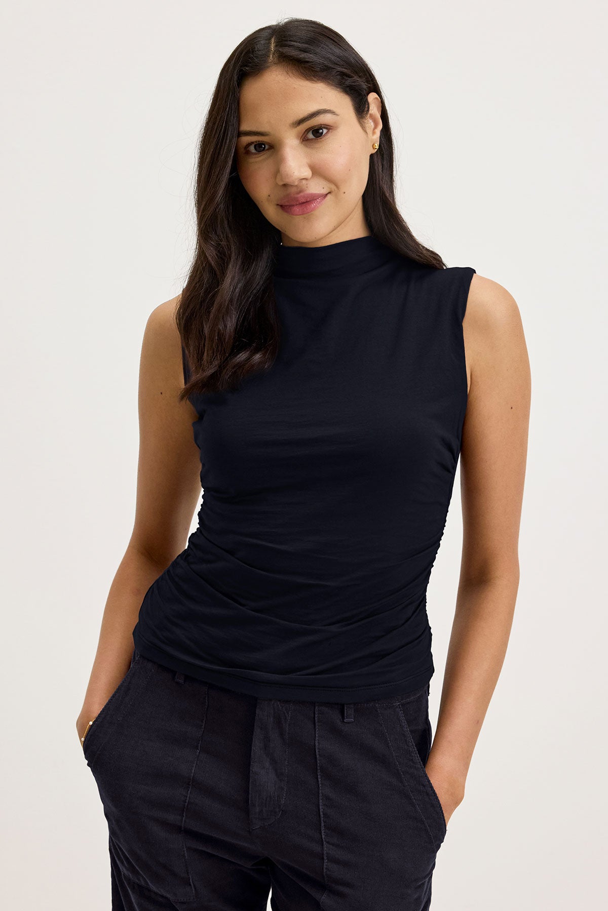   A person with long dark hair, wearing the Velvet by Graham & Spencer MADELINE TANK TOP made of gauzy whisper fabric and dark pants, stands with their hands in their pockets against a light background. 