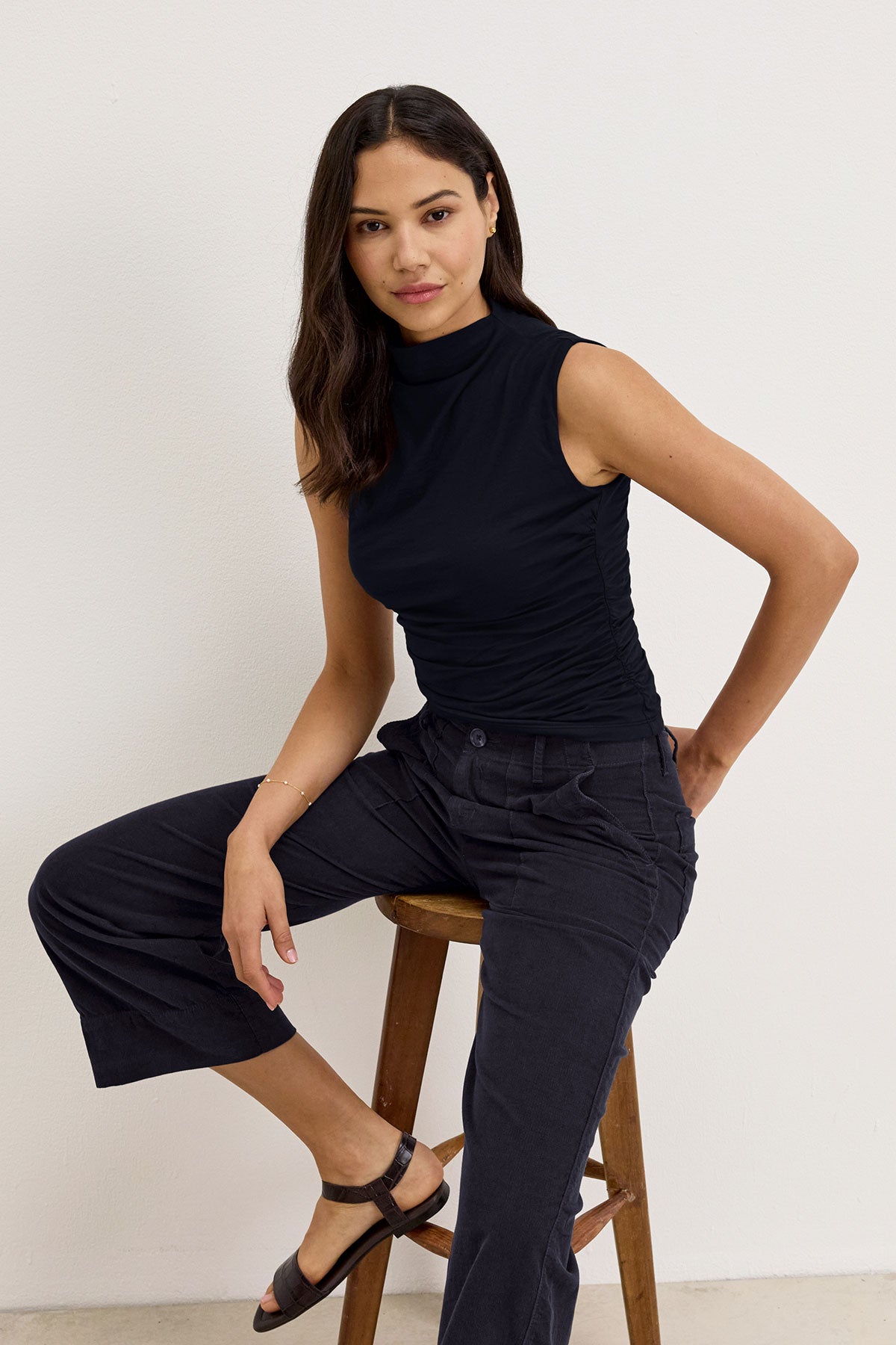   A woman sits on a stool against a plain background, wearing the MADELINE TANK TOP by Velvet by Graham & Spencer, paired with dark wide-leg pants and black sandals. She has long dark hair parted to the side. 