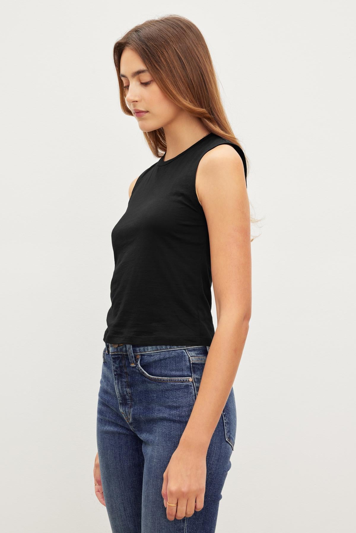 A woman wearing a MAGSON CREW NECK TANK TOP by Velvet by Graham & Spencer and blue jeans, standing side-profile against a plain background.-36418284454081