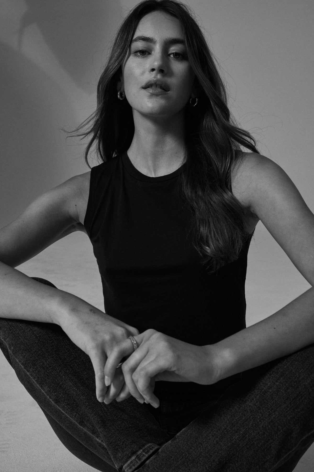 Black and white portrait of a woman in a Velvet by Graham & Spencer MAGSON CREW NECK TANK TOP and jeans sitting with crossed legs, looking at the camera.-36426074816705