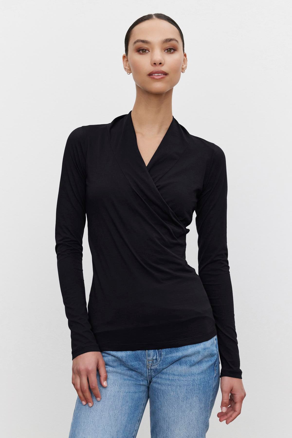   A person wearing the MERI TEE, a black long-sleeve wrap top with a V-neckline from Velvet by Graham & Spencer, and blue jeans stands against a plain white background. 