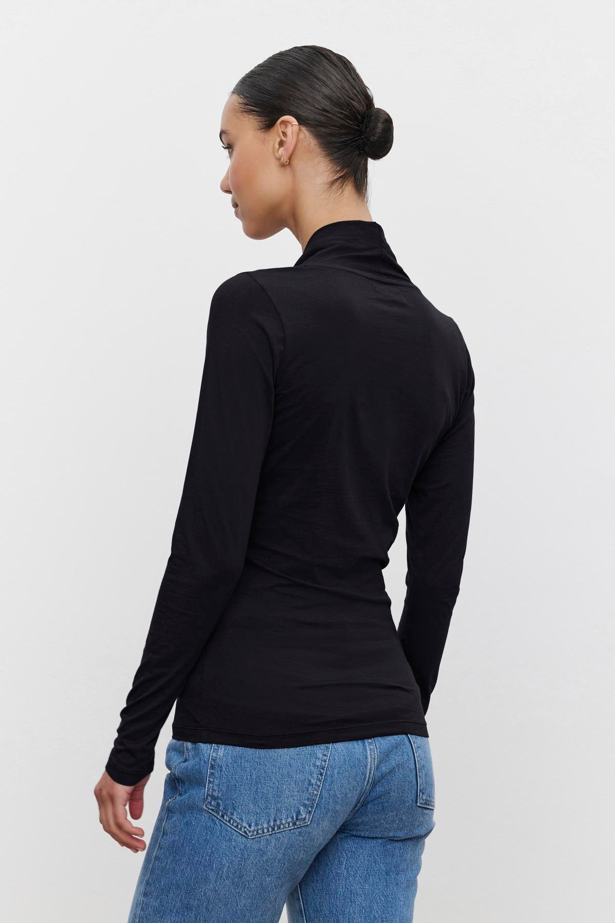   A person with tied-back hair stands facing away, wearing a black fitted MERI TEE by Velvet by Graham & Spencer and blue jeans against a plain white background. 