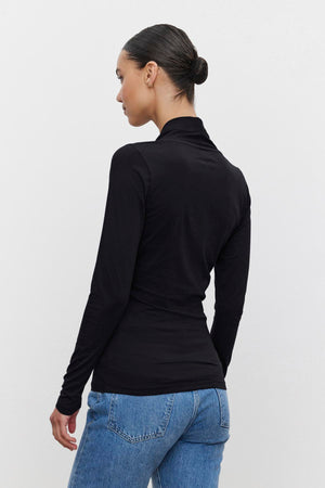 A person with tied-back hair stands facing away, wearing a black fitted MERI TEE by Velvet by Graham & Spencer and blue jeans against a plain white background.