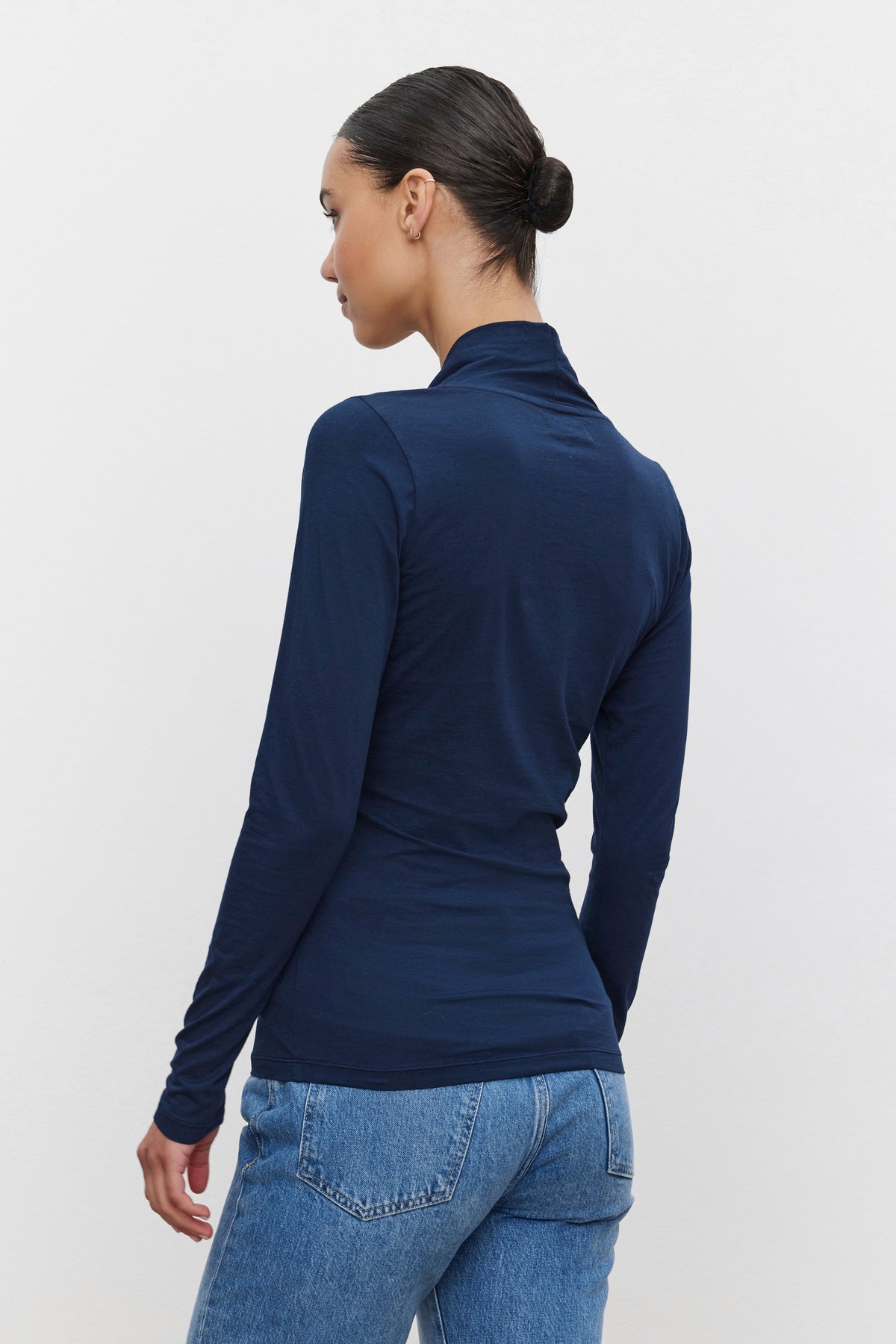   A person with dark hair is seen from behind, sporting a fitted silhouette in the MERI TEE long-sleeved, navy blue top by Velvet by Graham & Spencer and blue jeans. The hair is styled in a neat bun. 