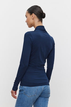 A person with dark hair is seen from behind, sporting a fitted silhouette in the MERI TEE long-sleeved, navy blue top by Velvet by Graham & Spencer and blue jeans. The hair is styled in a neat bun.