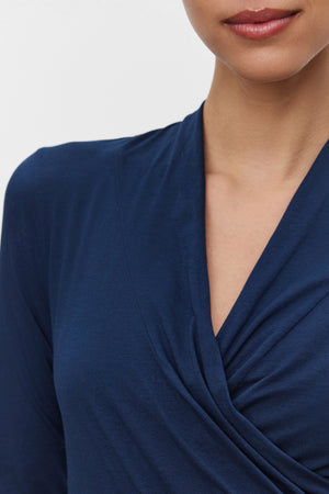 A person wearing the MERI TEE by Velvet by Graham & Spencer, a dark blue wrap-style top with a cross-over wrap detail, highlighting the V-neckline and upper shoulder.