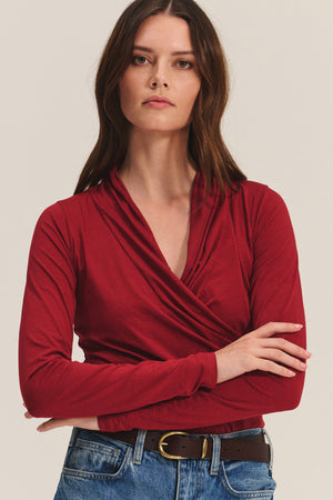 A woman with long brown hair wears the MERI TEE by Velvet by Graham & Spencer—a red wrap-detail V-neck tee—paired with blue jeans and a brown belt, standing with arms crossed against a plain background.