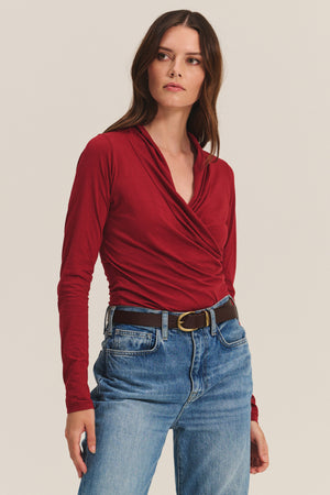 A woman wears the MERI TEE by Velvet by Graham & Spencer—a long-sleeve red wrap V-neck top—paired with blue jeans and a black belt, standing against a plain light background.