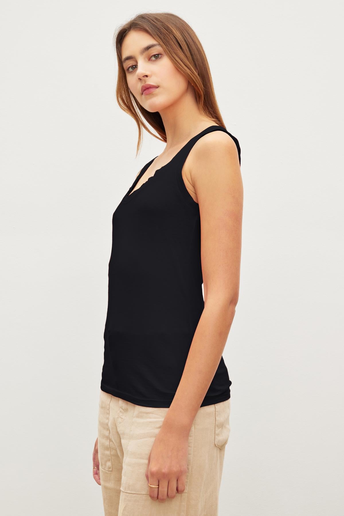   A woman in a Velvet by Graham & Spencer MOSSY GAUZY WHISPER FITTED TANK and beige pants stands against a plain background, looking at the camera with a neutral expression. 